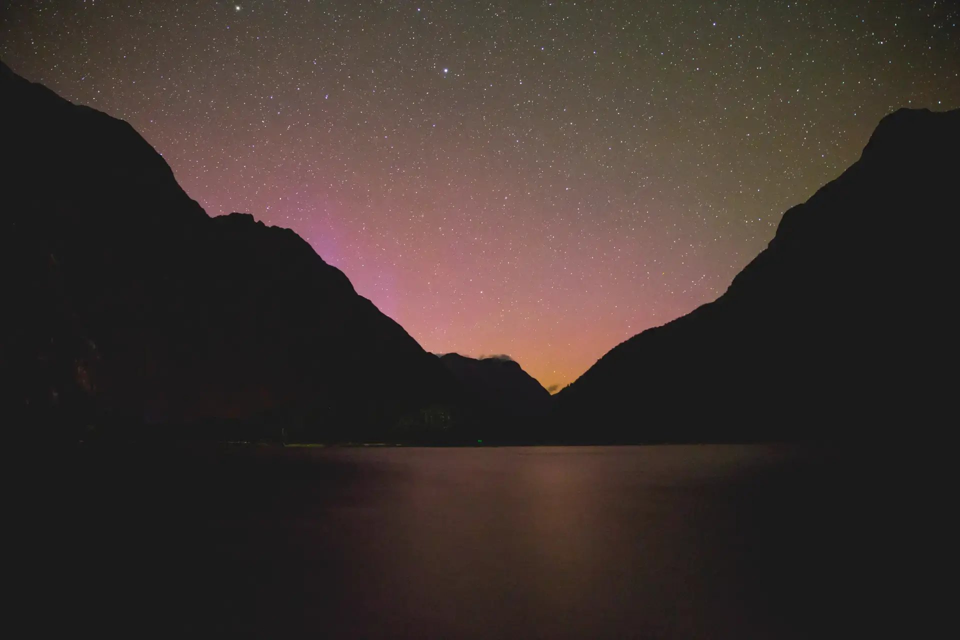 A night view in Milford Sound showing a dark silhouette of the fjord's mountains under a starry sky, with a faint purple and pink glow near the horizon.