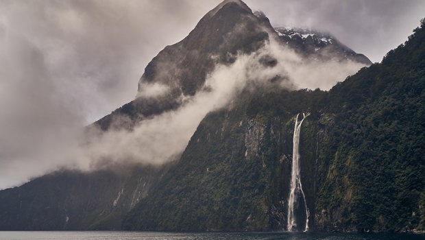 A cloudy day at Milford Sound, with a thin waterfall and mountain peaking through the clouds