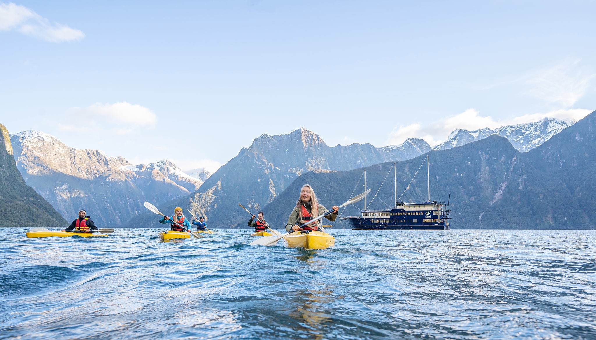 Group of friends kayaking in Milford Sound with boat and mountains in the background