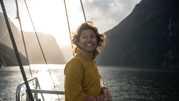 A woman smiles on the front deck of a vessel cruising Milford Sound, with the sun setting behind