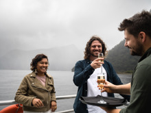 A couple accept a glass of champaign each from a server on an open deck of a vessel while cruising Milford Sound