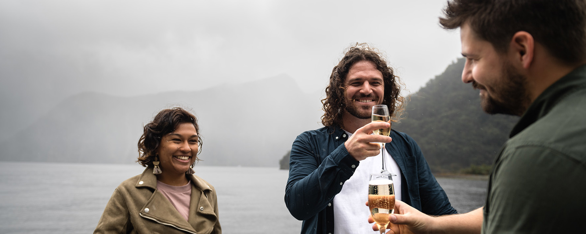 A couple accept a glass of champaign each from a server on an open deck of a vessel while cruising Milford Sound