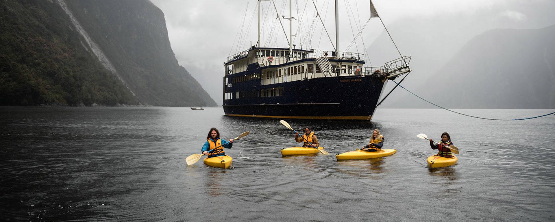 A group of four kayakers floating on Milford Sound in front of the Milford Wanderer vessel