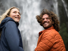 A couple look back over their shoulder with a cascading waterfall behind them