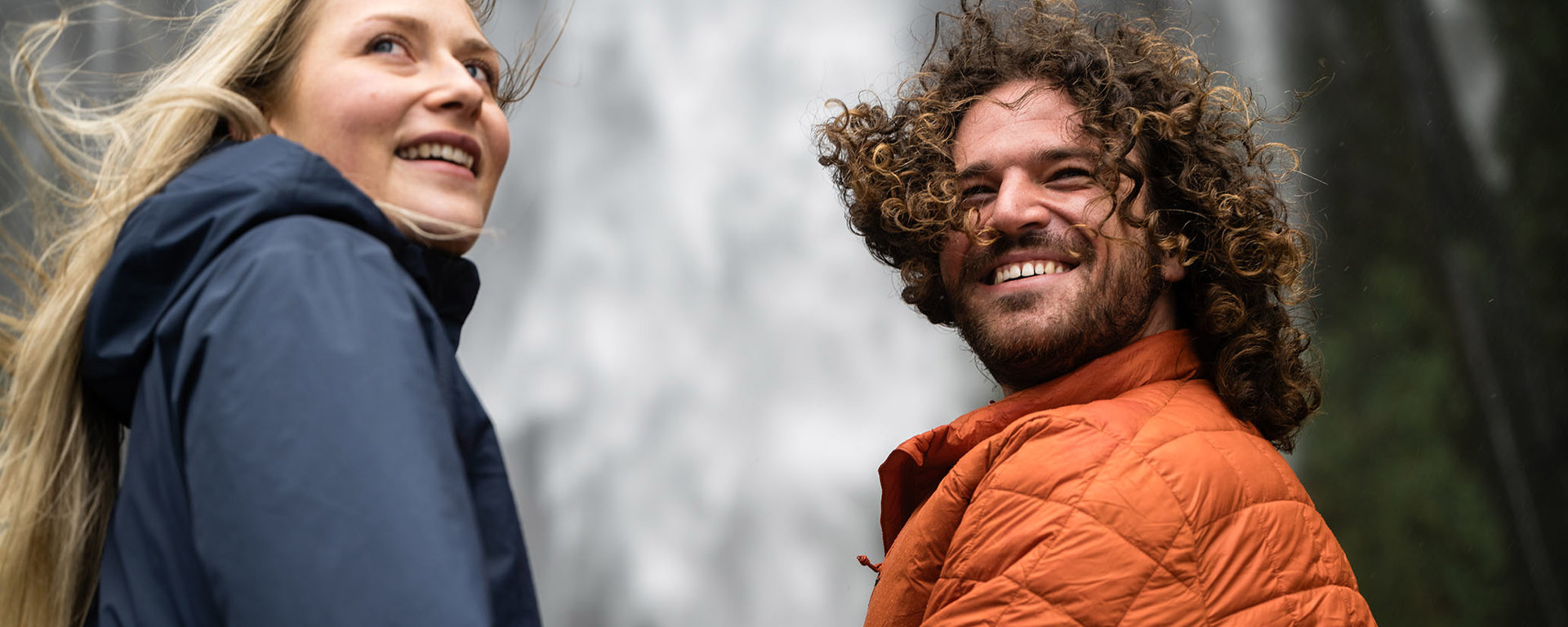 A couple look back over their shoulder with a cascading waterfall behind them