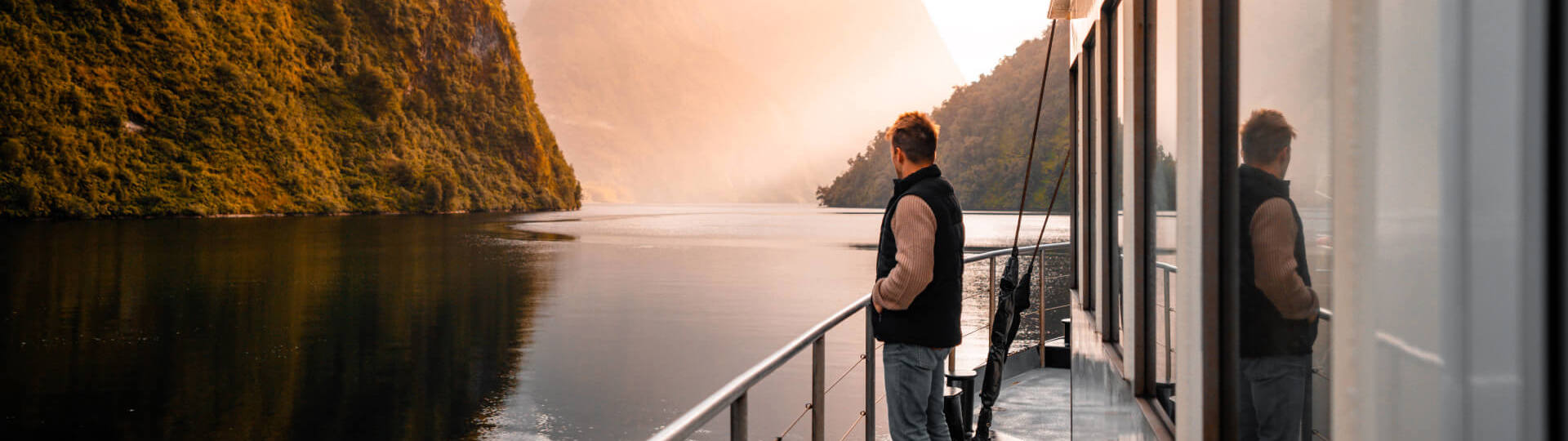 A man stands on the side deck of a vessel cruising through Doubtful  Sound at golden hour