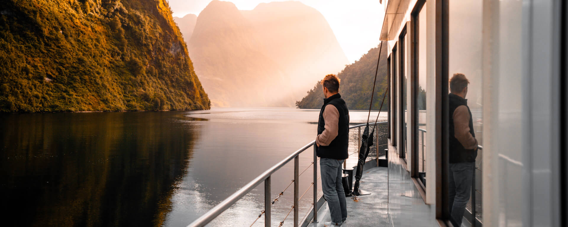 A man stands on the side deck of a vessel cruising through Doubtful  Sound at golden hour