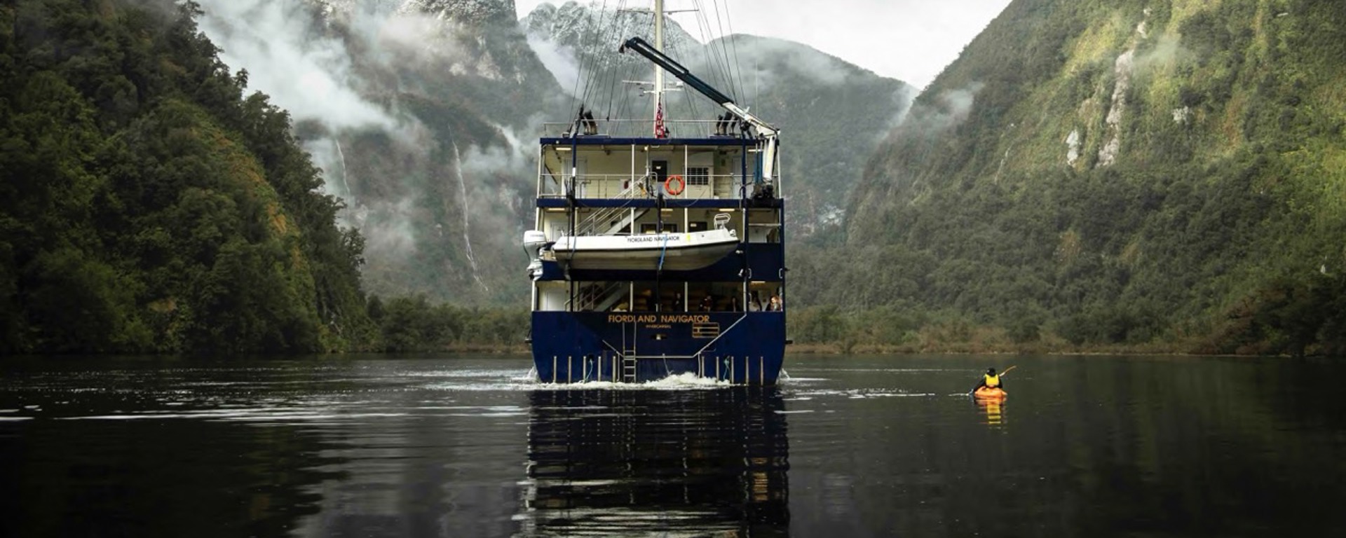 A kayaker paddles next the Fiordland Navigator vessel on Doubtful Sound, surrounded by snowy mountains