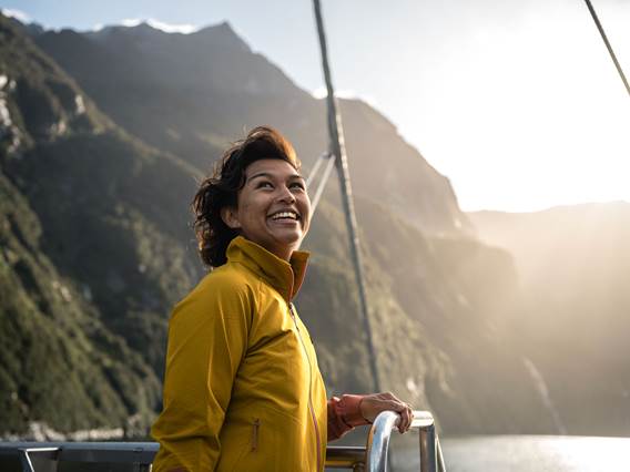 A woman in a bright yellow jacket enjoys the view of sunny Doubtful Sound from the top deck of a vessel