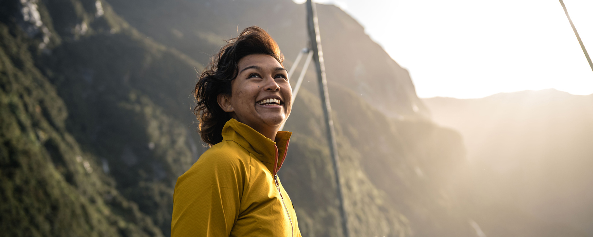 A woman in a bright yellow jacket enjoys the view of sunny Doubtful Sound from the top deck of a vessel