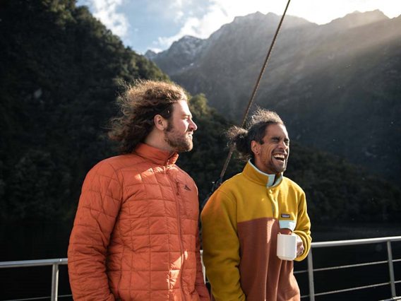 Two men enjoy a hot drink on the outside deck onboard a cruise on Doubtful Sound