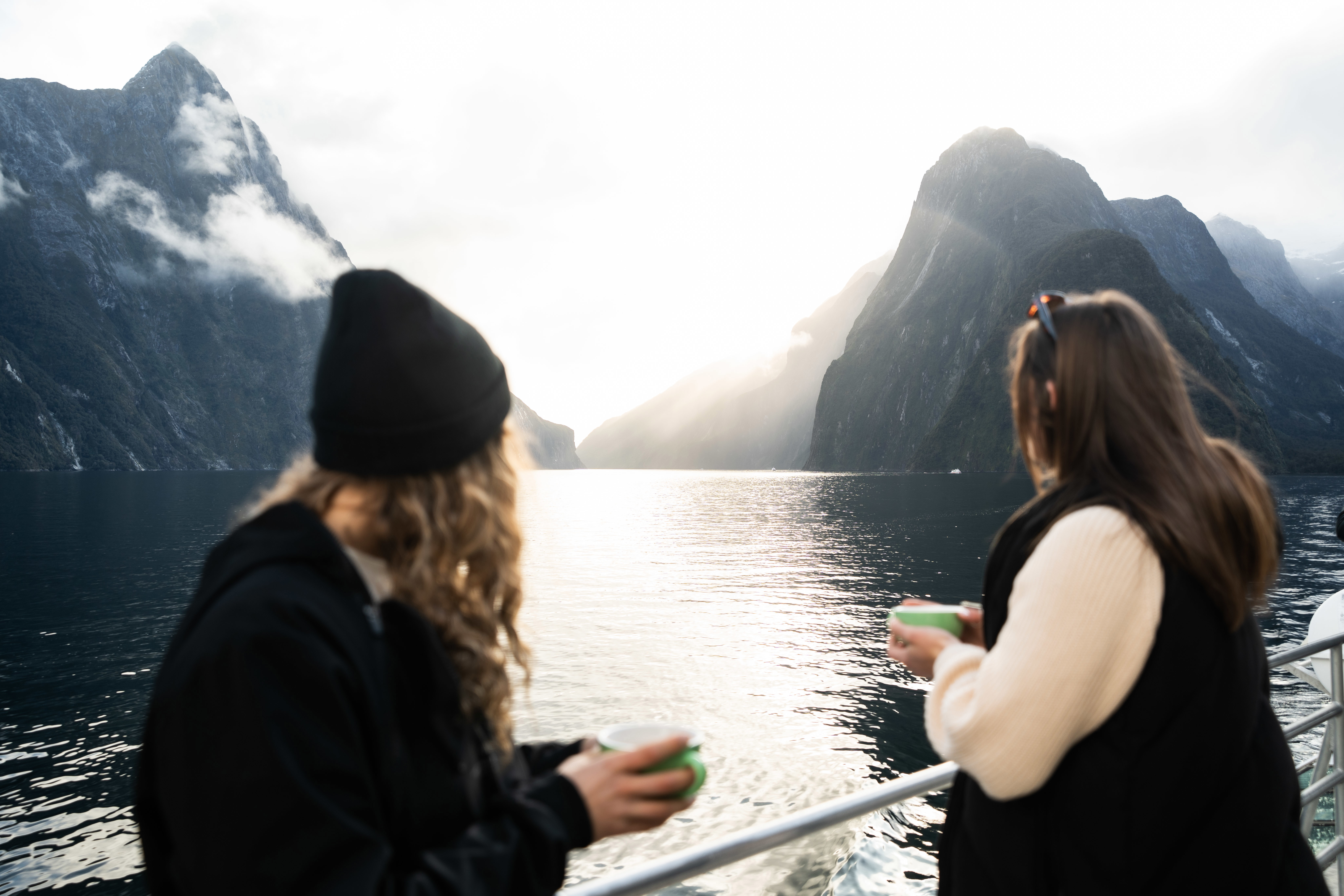 Two women standing outside on a vessel lean against the barrier holding mugs and looking out to the view of spectacular mountains. 