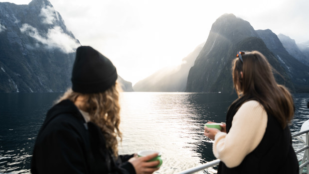 Two women standing outside on a vessel lean against the barrier holding mugs and looking out to the view of spectacular mountains. 