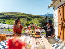 Two people toast with wine glasses at a rustic outdoor table, surrounded by scenic green hills and a sunny countryside.