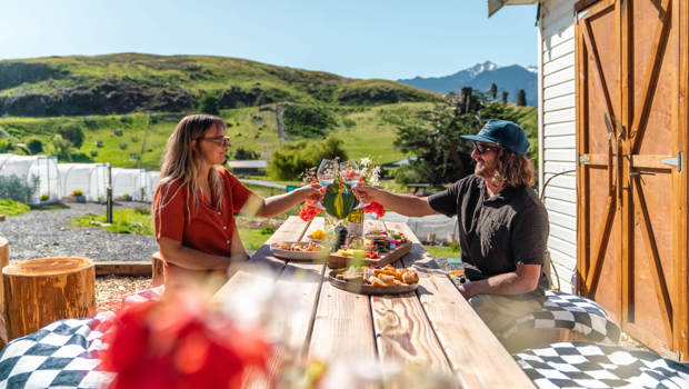 Two people toast with wine glasses at a rustic outdoor table, surrounded by scenic green hills and a sunny countryside.