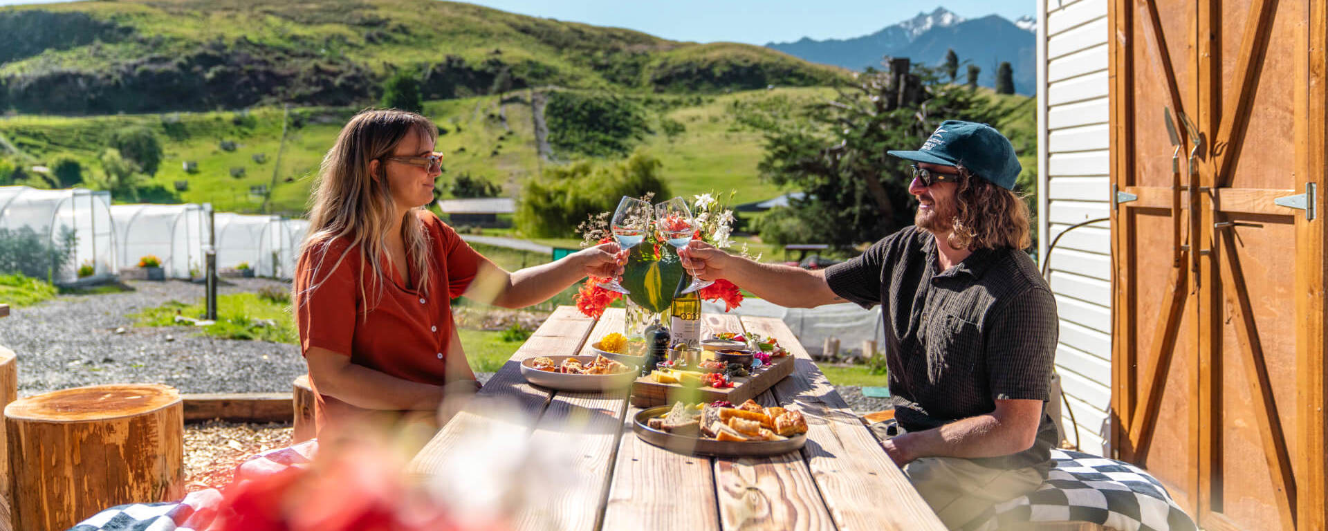 Two people toast with wine glasses at a rustic outdoor table, surrounded by scenic green hills and a sunny countryside.