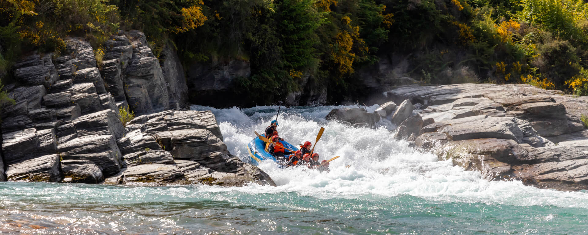 Rafting group tackling white-water rapids surrounded by rocky cliffs and vibrant greenery on the Shotover River in New Zealand.