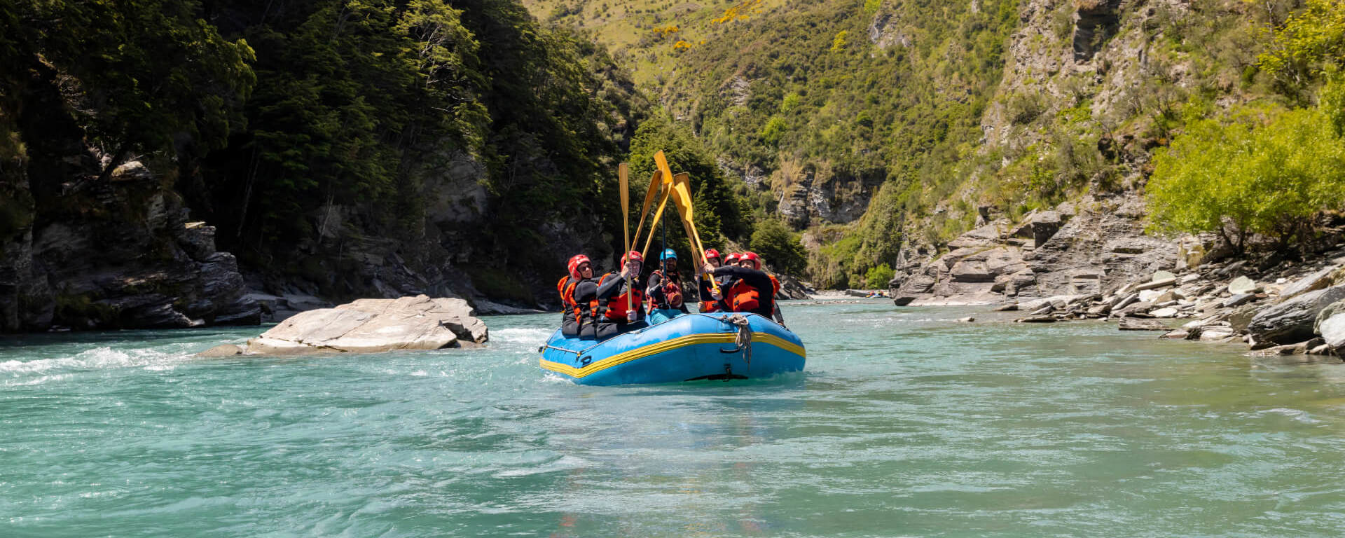 Rafters celebrating with paddles raised high while drifting through a serene canyon on the turquoise waters of the Shotover River.