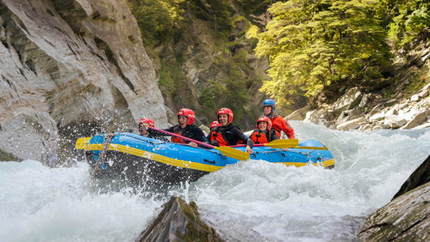 Thrilled rafters splashing through fast-moving rapids surrounded by dramatic cliffs and lush greenery on the Shotover River.