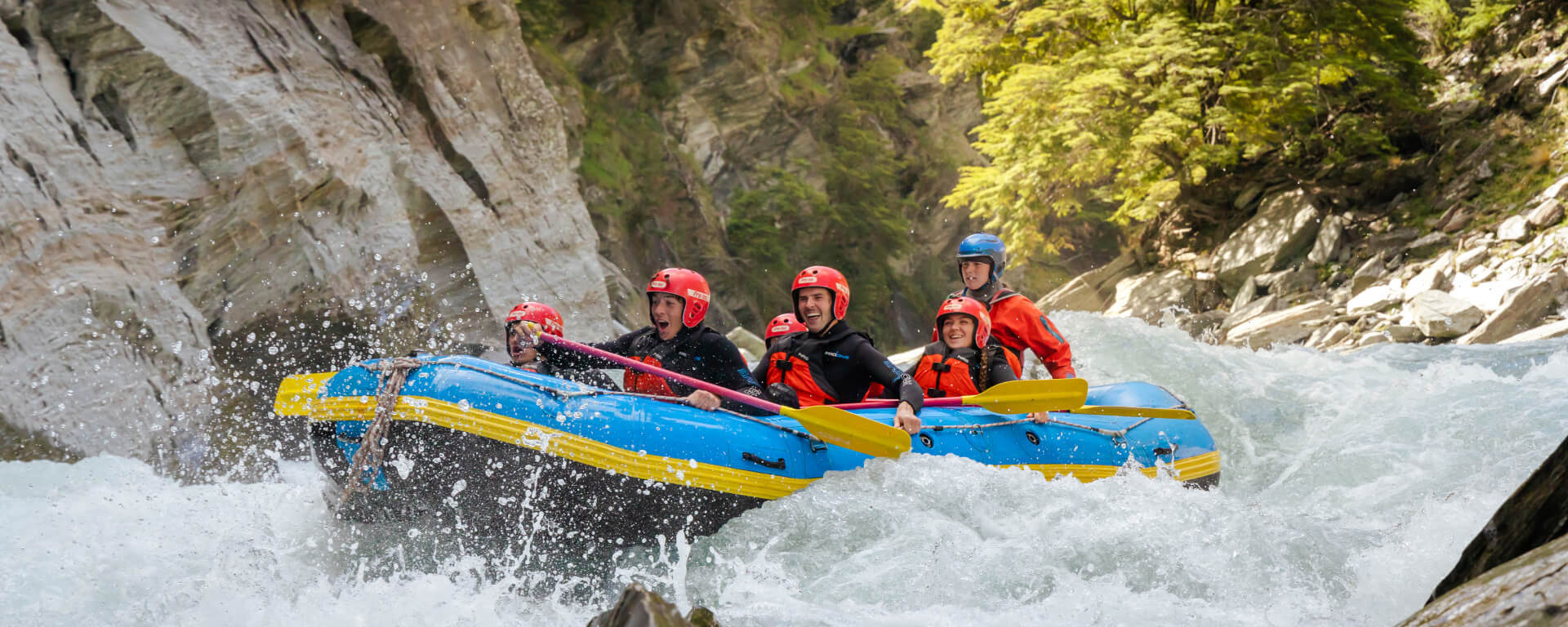 Thrilled rafters splashing through fast-moving rapids surrounded by dramatic cliffs and lush greenery on the Shotover River.