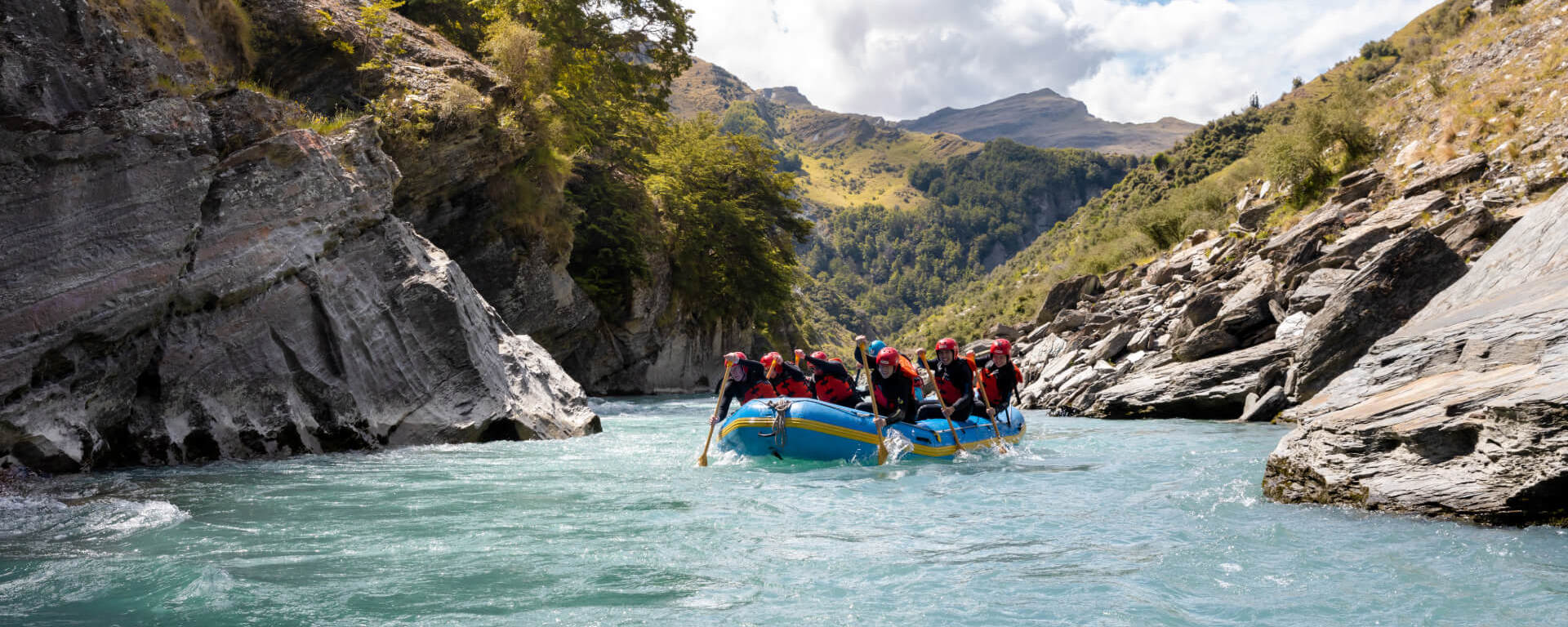 Rafting group paddling through turquoise waters in a scenic canyon surrounded by rocky cliffs and lush greenery on the Shotover River.