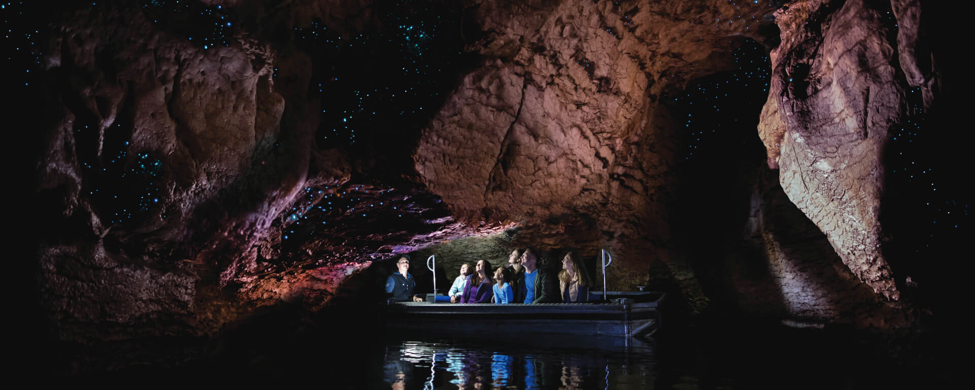 A small boat glides through the Te Anau glowworm caves, where visitors gaze at thousands of glowing blue lights on the cave ceiling, reflecting off calm waters.