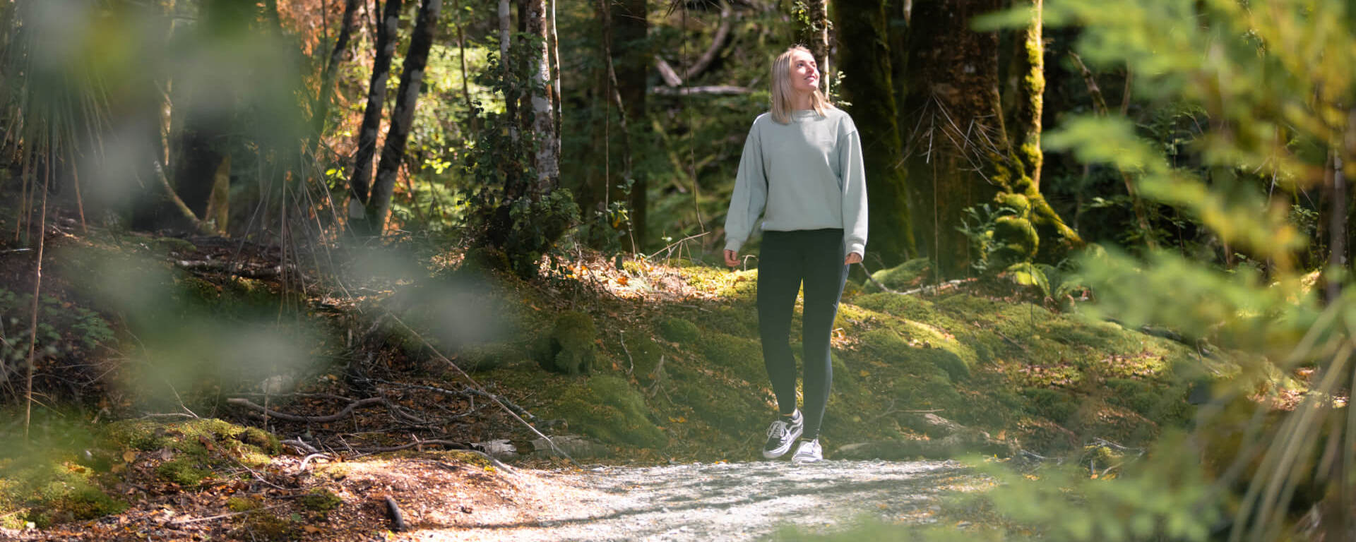 A woman walks along a forest path surrounded by lush greenery and dappled sunlight, enjoying the serene natural beauty near the Te Anau Glowworm Caves.