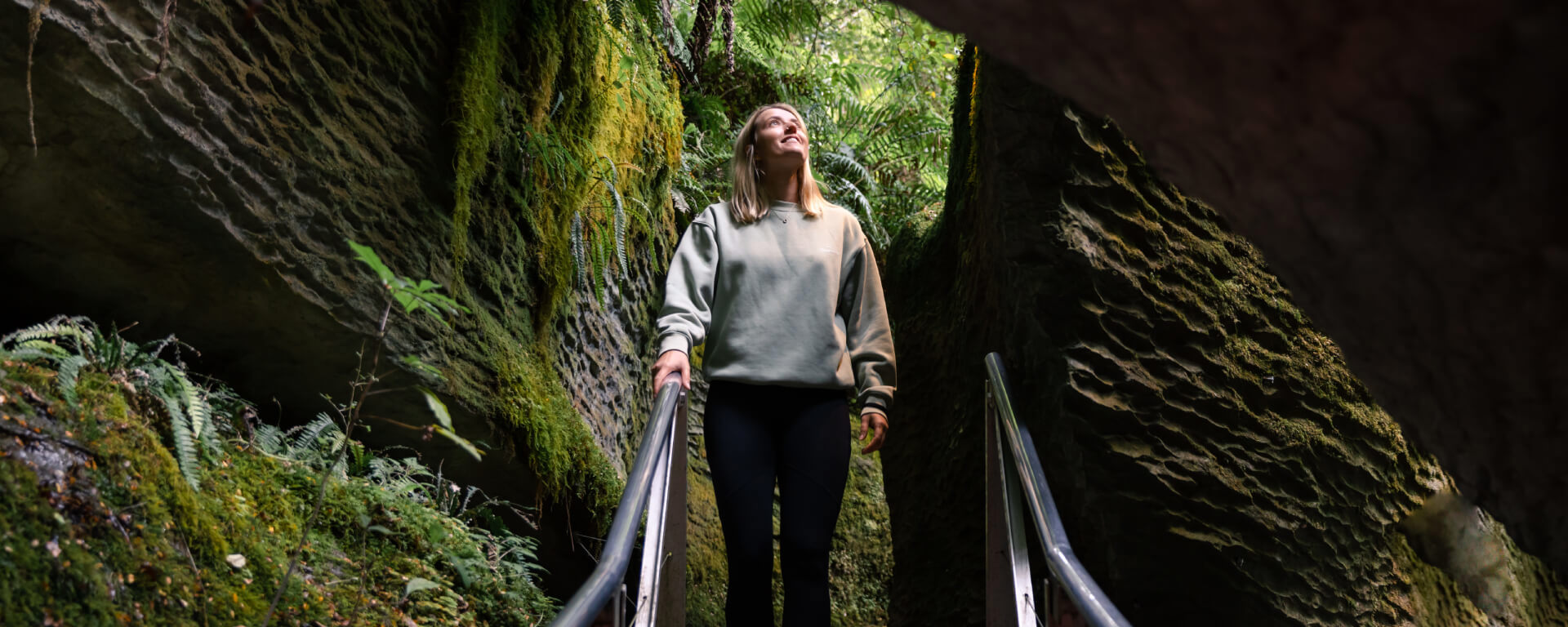 A woman stands on a walkway surrounded by moss-covered rock walls, gazing upward as she enters the lush and enchanting entrance to the Te Anau Glowworm Caves.