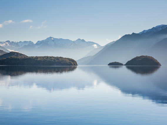  A tranquil lake reflects forested islands and snow-capped mountains under a clear blue sky, creating a stunning backdrop on the start of a Te Anau glowworm caves tour.