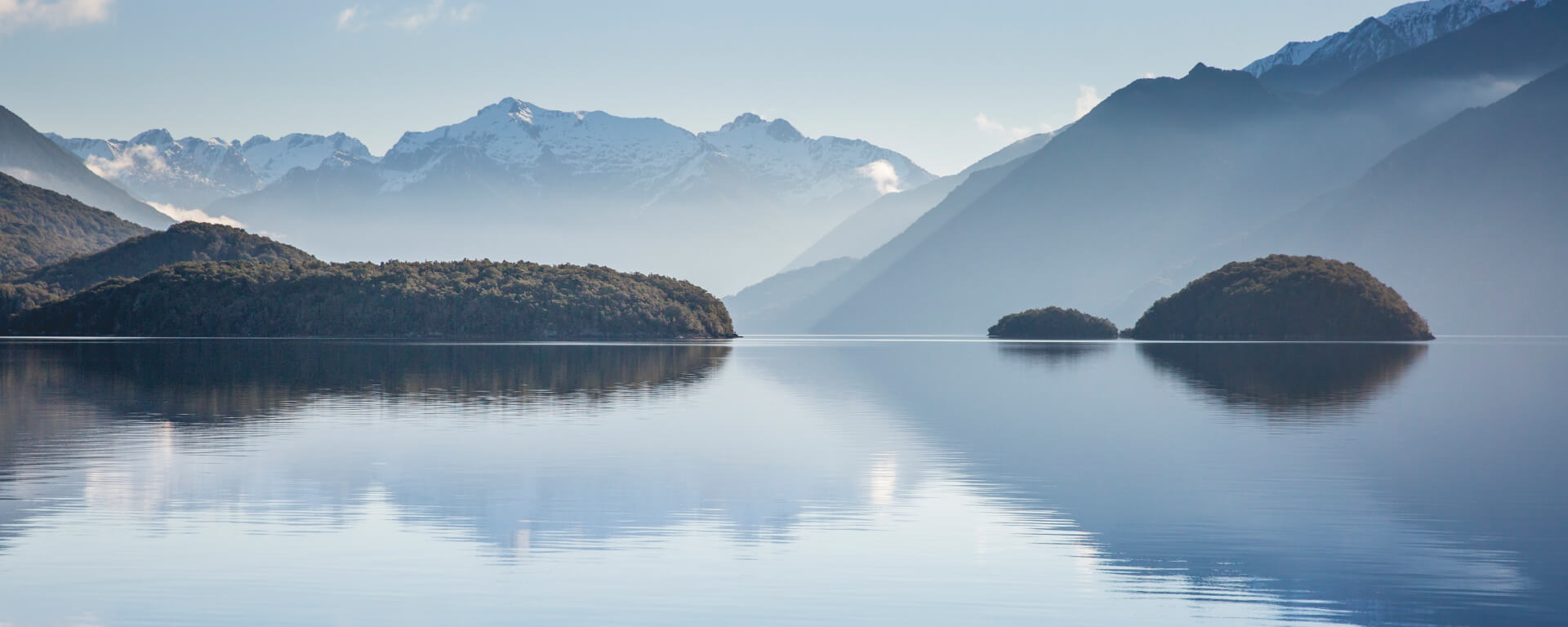  A tranquil lake reflects forested islands and snow-capped mountains under a clear blue sky, creating a stunning backdrop on the start of a Te Anau glowworm caves tour.