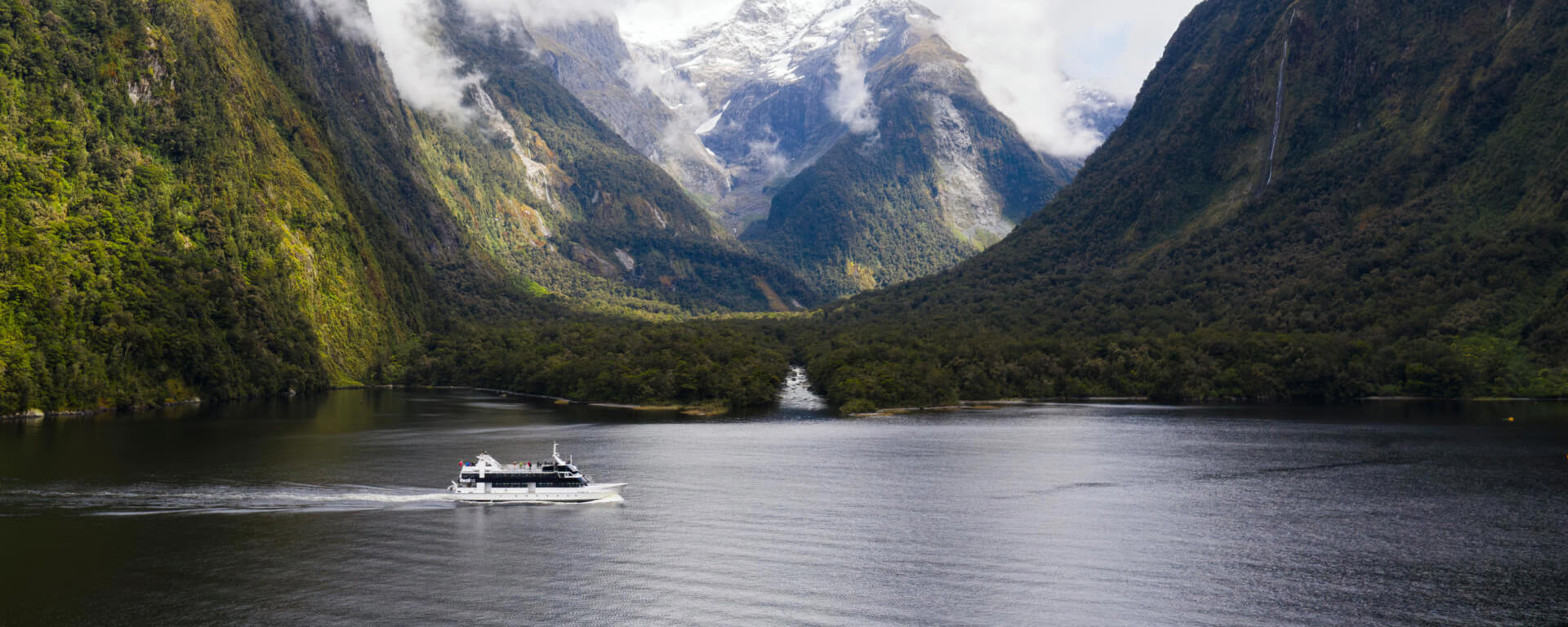 Wide shot of a vessel cruising in Milford Sound with snow covered mountains in the background