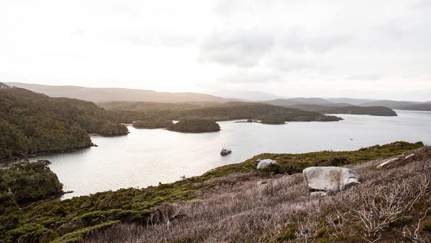 Expansive view from Bald Cone on Stewart Island, showcasing rugged coastline, scattered forested islands, and a boat cruising in the calm, reflective waters below.