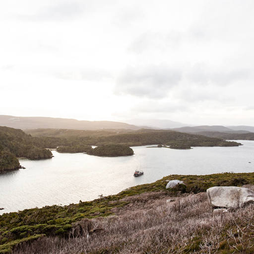 Expansive view from Bald Cone on Stewart Island, showcasing rugged coastline, scattered forested islands, and a boat cruising in the calm, reflective waters below.