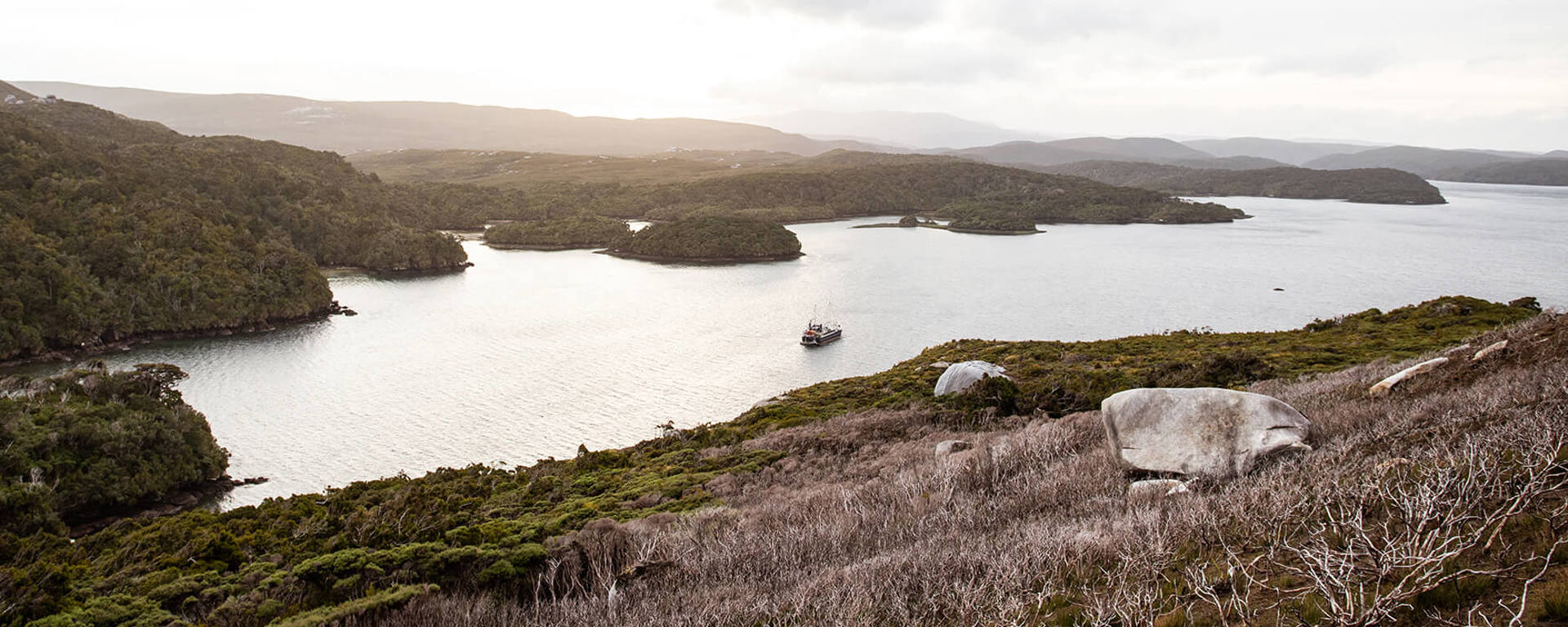 Expansive view from Bald Cone on Stewart Island, showcasing rugged coastline, scattered forested islands, and a boat cruising in the calm, reflective waters below.