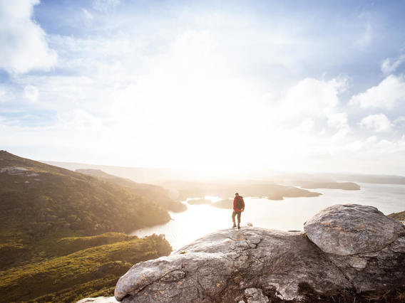 Man standing on a rocky outcrop atop Bald Cone, Stewart Island, overlooking a stunning landscape of rolling hills, shimmering water, and a bright, expansive sky.