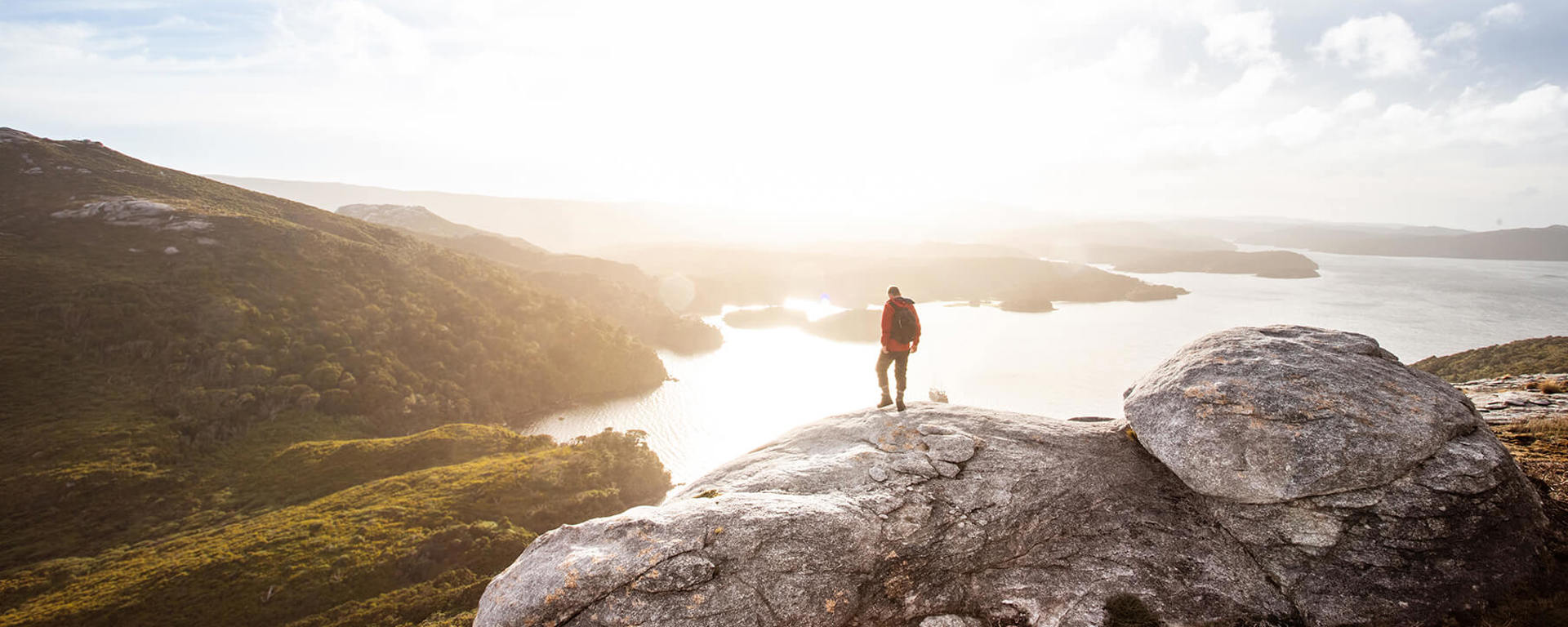 Man standing on a rocky outcrop atop Bald Cone, Stewart Island, overlooking a stunning landscape of rolling hills, shimmering water, and a bright, expansive sky.