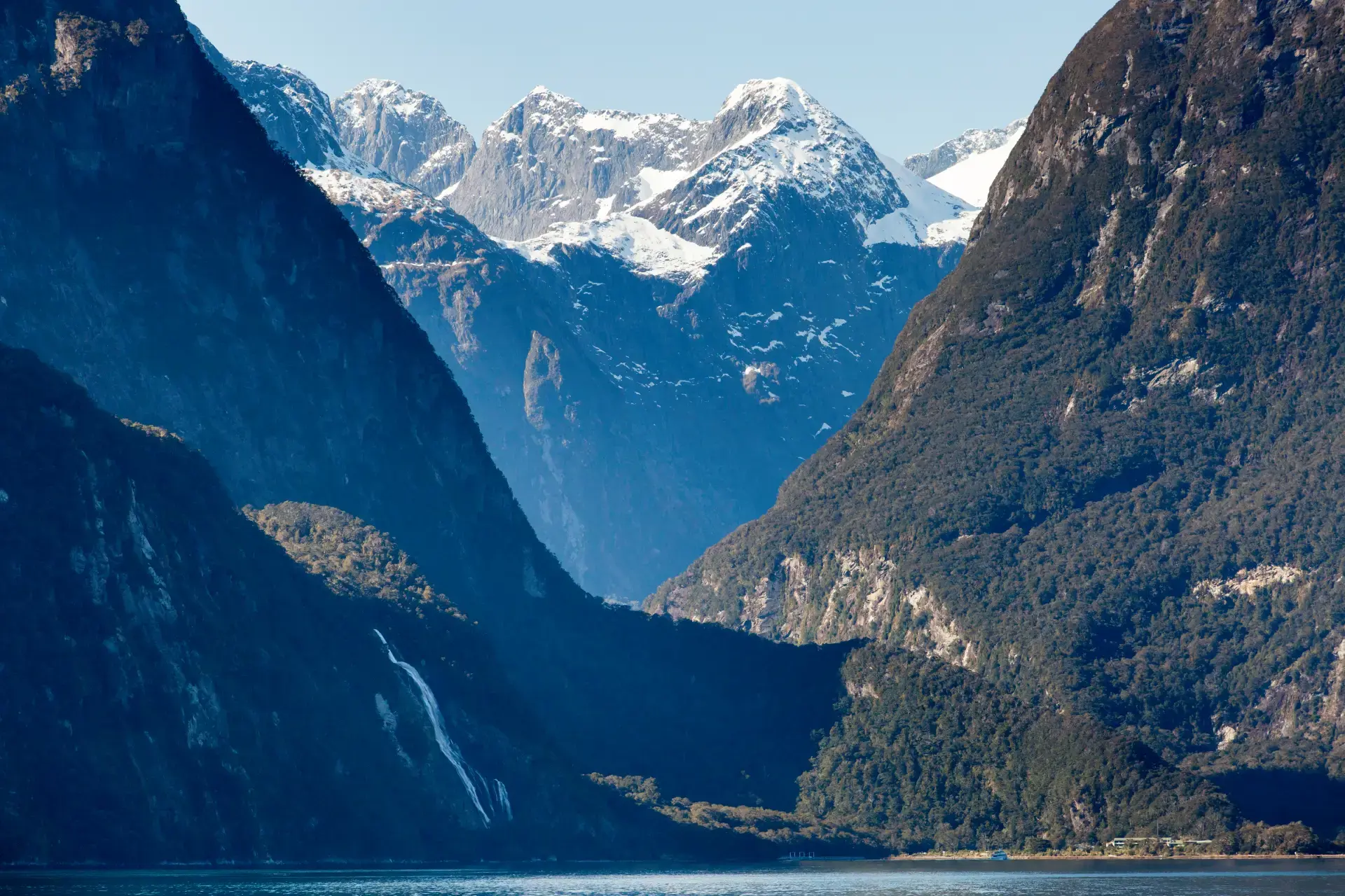 Snow-capped peaks towering over a deep fiord in Milford Sound with Stirling Falls waterfall gushing in the foreground. 