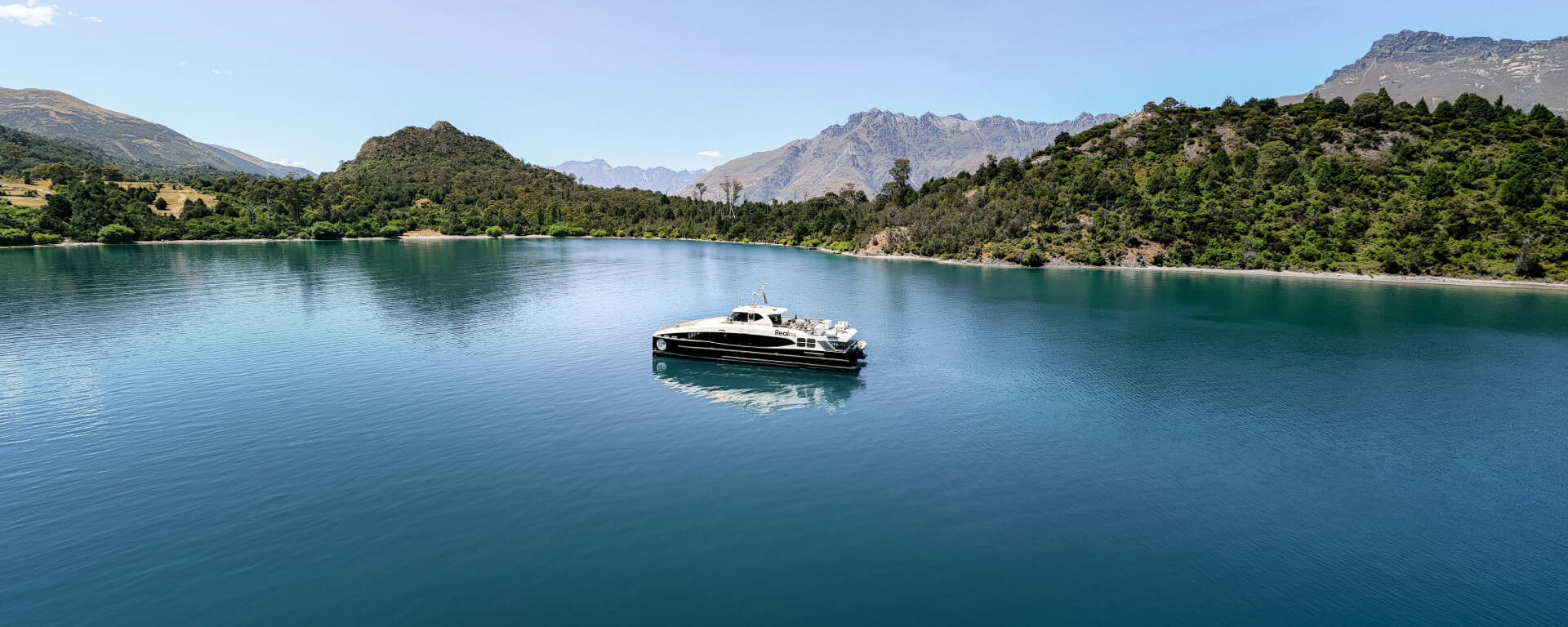 Spirit of Queenstown cruise boat on the tranquil waters of Bob’s Cove, surrounded by lush greenery and mountain landscapes under a clear blue sky.