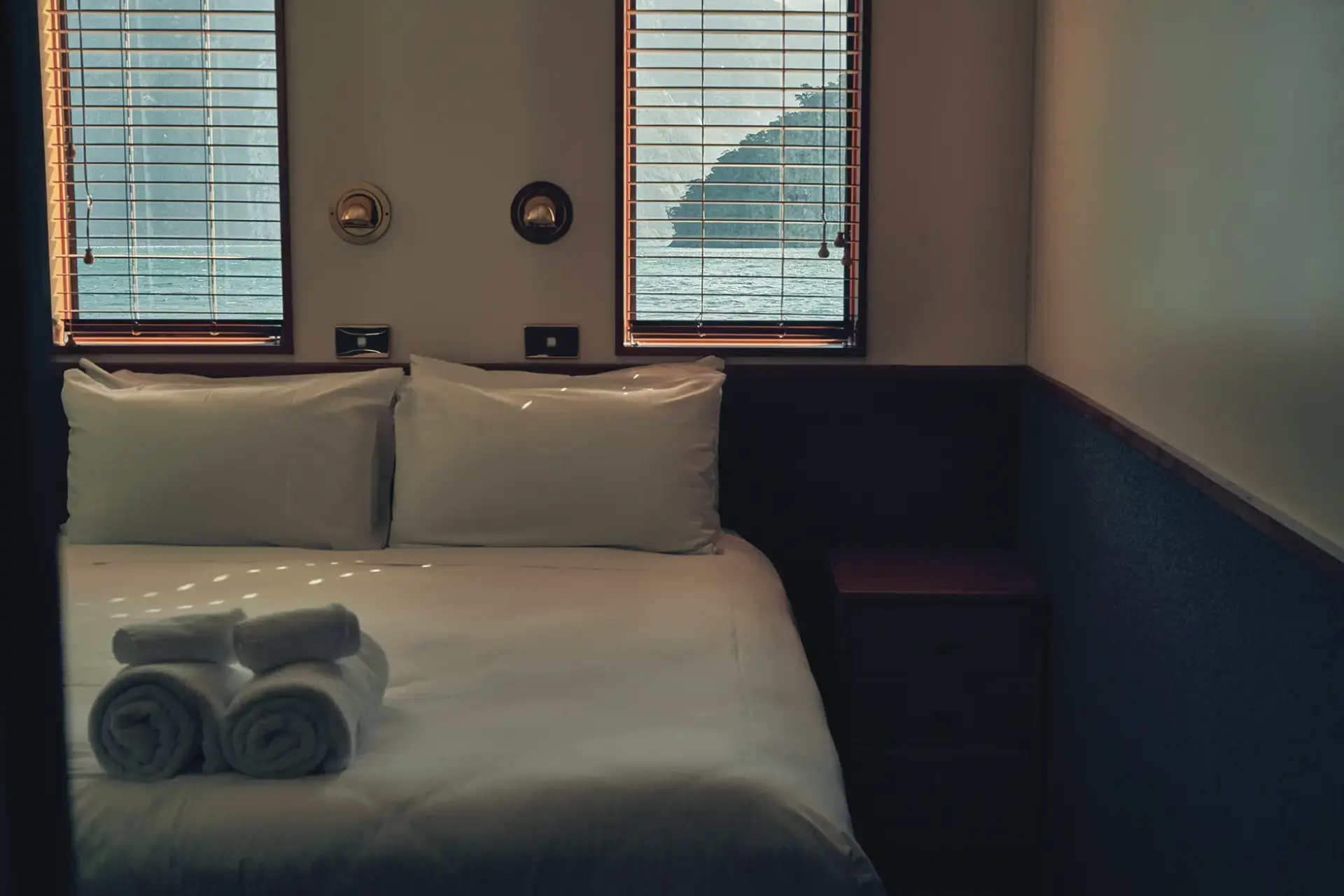 The interior of a cozy cabin on the Milford Sound Overnight Cruise, featuring a neatly made bed with soft lighting, a window overlooking the water, and wooden accents.