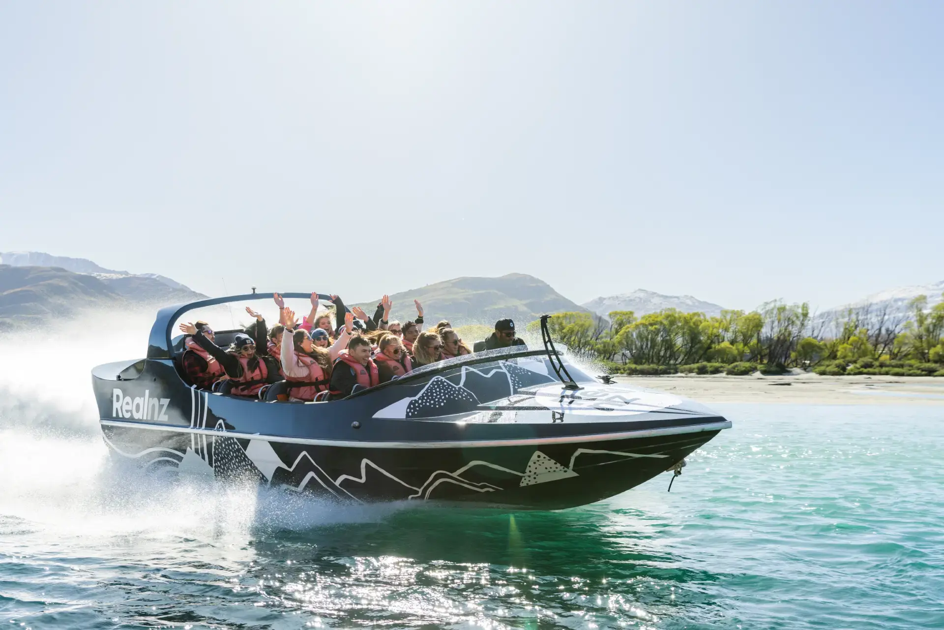 A RealNZ jet boat spins in the water on Lake Whakatipu on a clear blue day, full of guests smiling and raising their arms in excitement. 