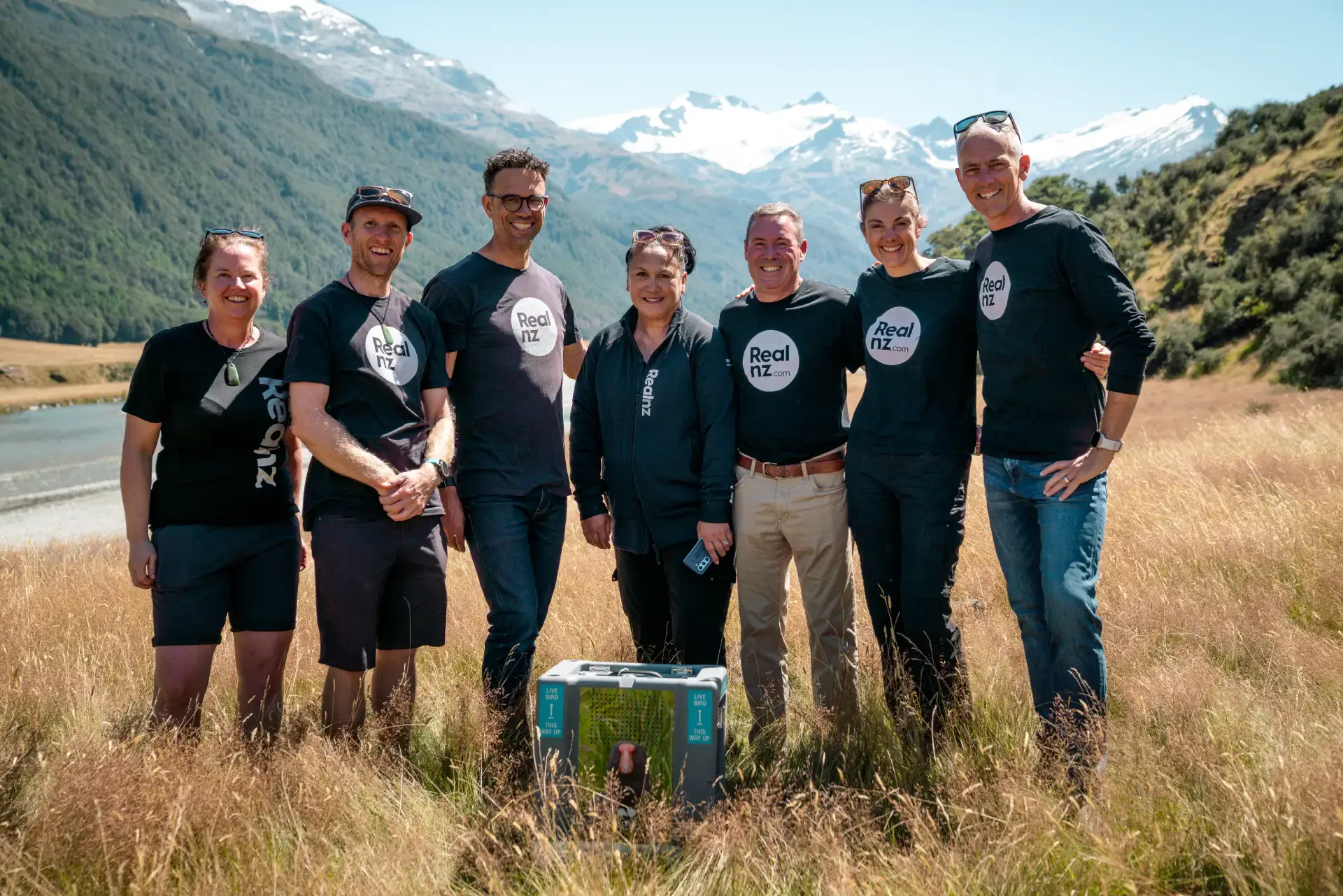 A smiling RealNZ team poses in Rees Valley with a takahē transport crate in front of them, surrounded by tall grass and snow-capped mountains in the background.