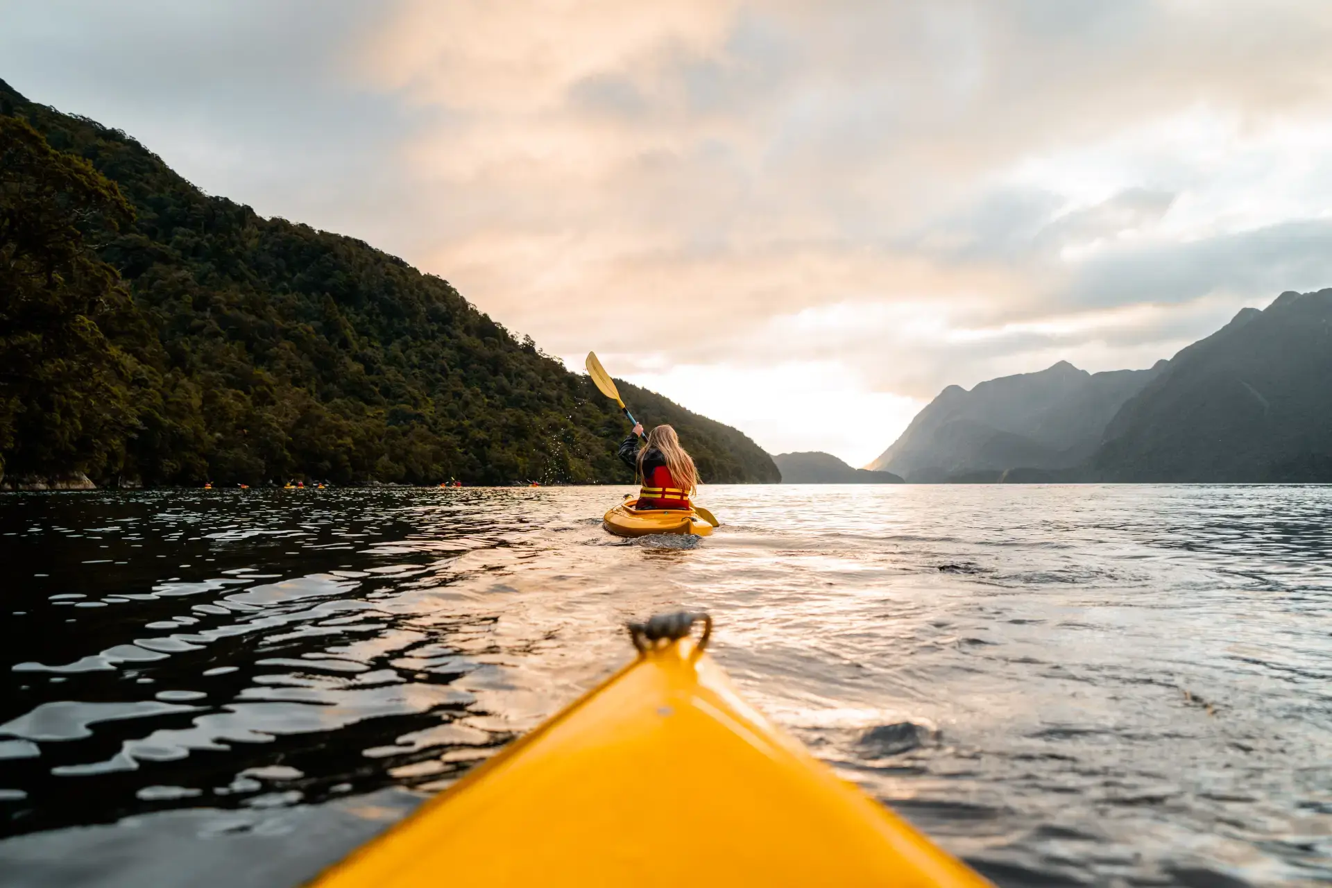 A kayaker paddling through Milford Sound at sunset, surrounded by mountains.