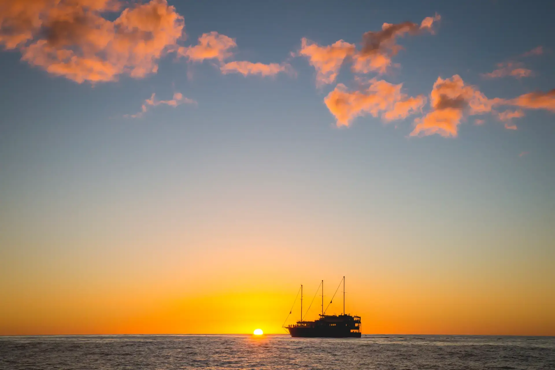 The Milford Mariner boat cruises through Milford Sound at sunset, with the sky glowing orange and pink over calm waters, and silhouettes of mountains in the background.