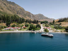 Spirit of Queenstown cruise boat docked at Walter Peak High Country Farm, surrounded by lush greenery, red-roofed buildings, and dramatic mountain scenery.