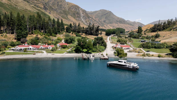 Spirit of Queenstown cruise boat docked at Walter Peak High Country Farm, surrounded by lush greenery, red-roofed buildings, and dramatic mountain scenery.