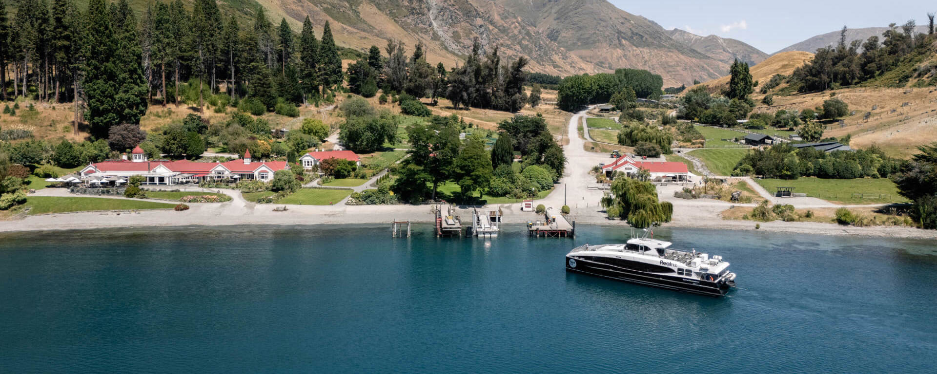 Spirit of Queenstown cruise boat docked at Walter Peak High Country Farm, surrounded by lush greenery, red-roofed buildings, and dramatic mountain scenery.