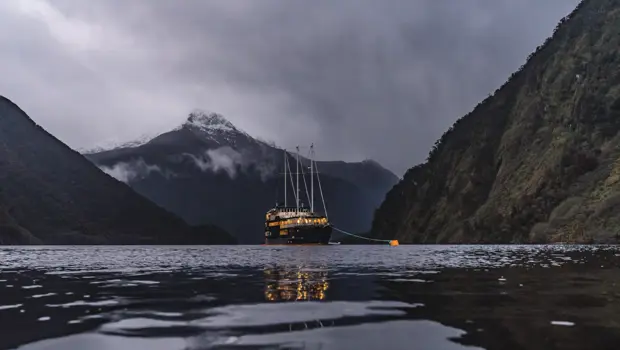 The Milford Mariner overnight cruise boat anchors in Milford Sound for the evening, surrounded by darkening waters and lit orange from the lights within. 