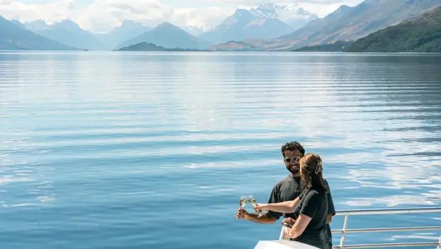 A couple cheers two glasses of champagne on the Queenstown Lake Cruise, while looking at the crystal clear water on a clear blue day.